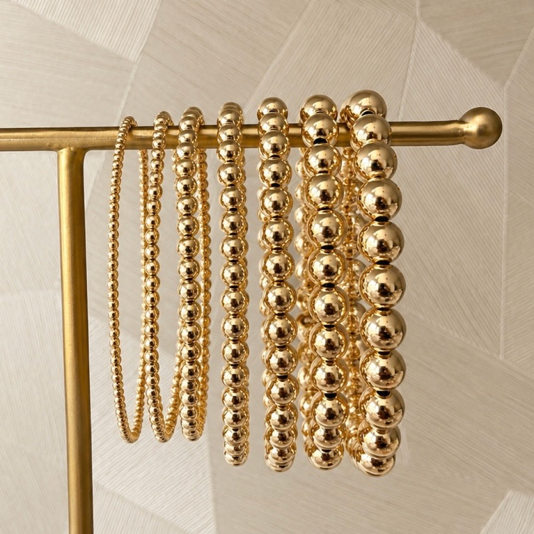 Zeven gouden kralenarmbanden van gold filled 14K in oplopende kraalmaten aan een gouden sieradenrek, van Bedels & Bedels.