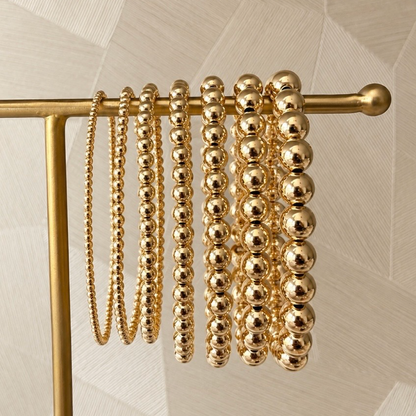 Zeven gouden kralenarmbanden van gold filled 14K in oplopende kraalmaten aan een gouden sieradenrek, van Bedels & Bedels.